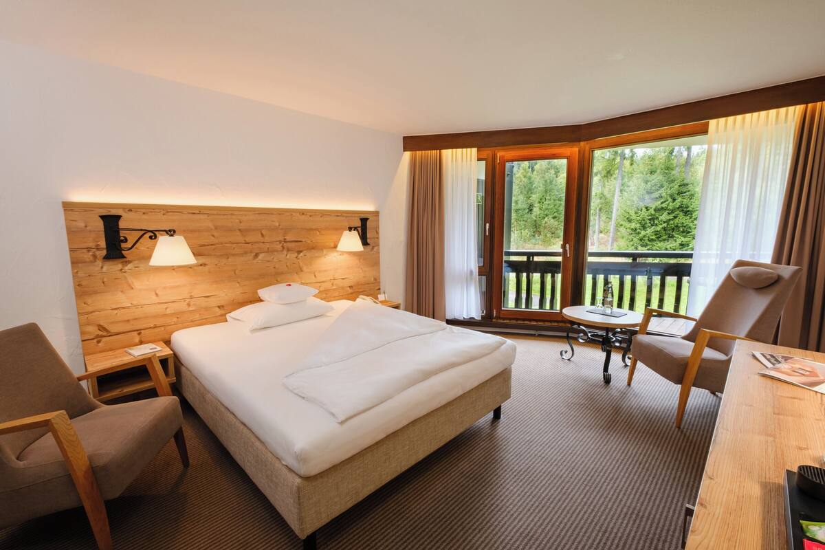 A hotel room with a bed, two bedside lamps, a wooden headboard, a desk with a chair, a small table, and a balcony with curtains.