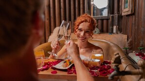 A person toasting with a glass at a wooden table with rose petals, surrounded by various drinks and a candle.