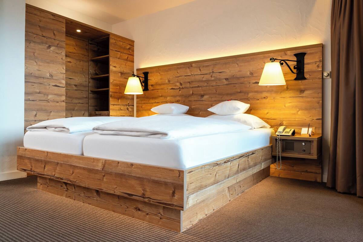 A wooden-framed bed with white linens, two bedside lamps, a bedside telephone, and a small wooden nightstand.
