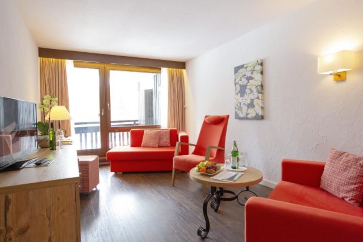 Una habitación de hotel moderna con un sofá rojo, un escritorio de madera, una pequeña mesa redonda con frutas y bebidas, y un ventanal con cortinas.