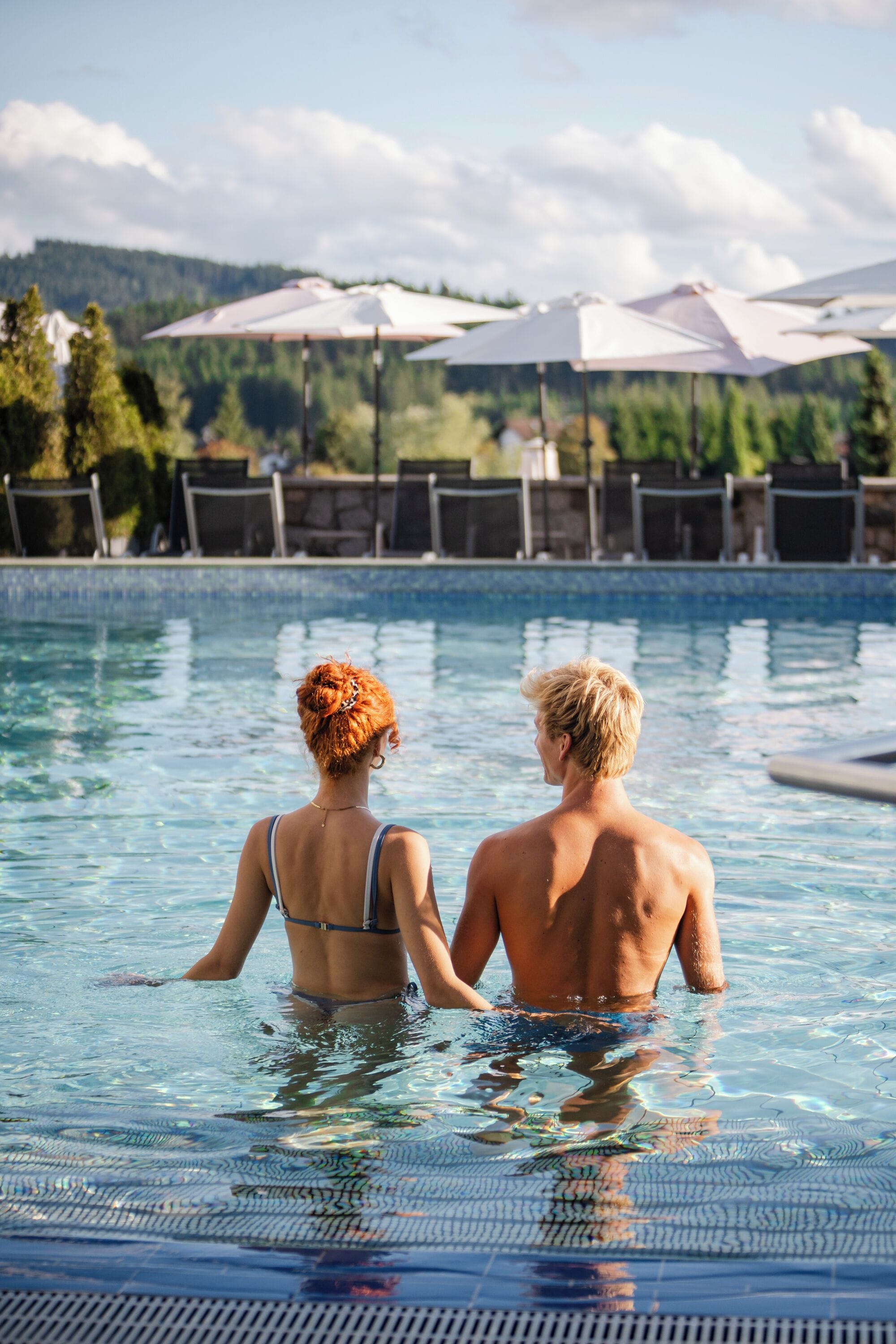 Ein Schwimmbecken mit zwei Personen im Wasser, umgeben von Liegestühlen und Sonnenschirmen.