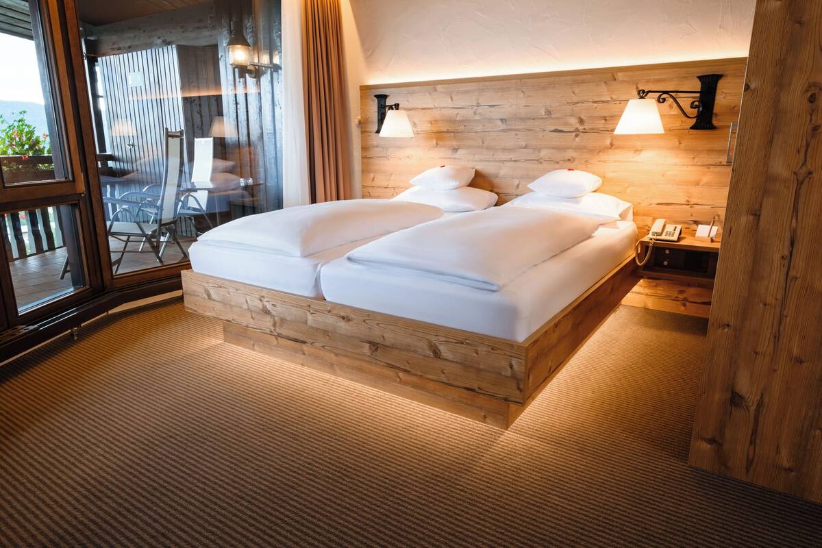 A double bed with a wooden headboard, a bedside table with a lamp, and a view through a window.