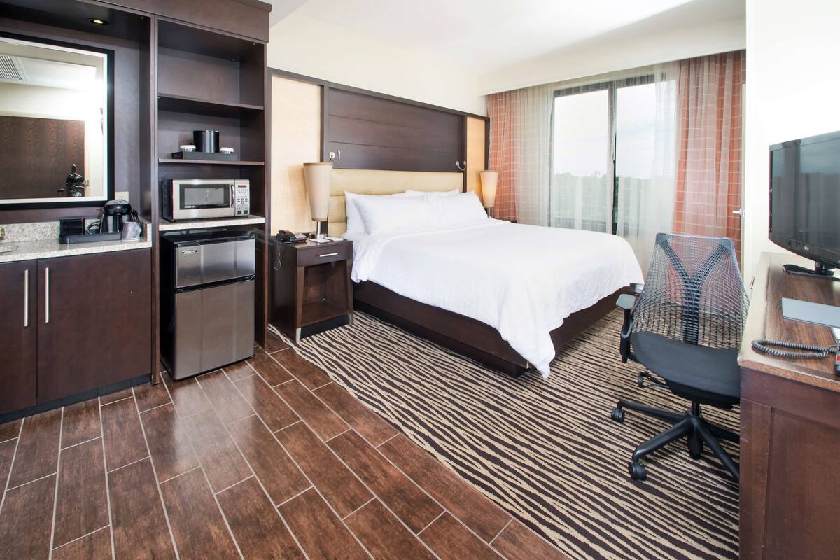 A hotel room with a bed, a desk with a chair, a microwave, a coffee maker, a mini-fridge, and a TV.