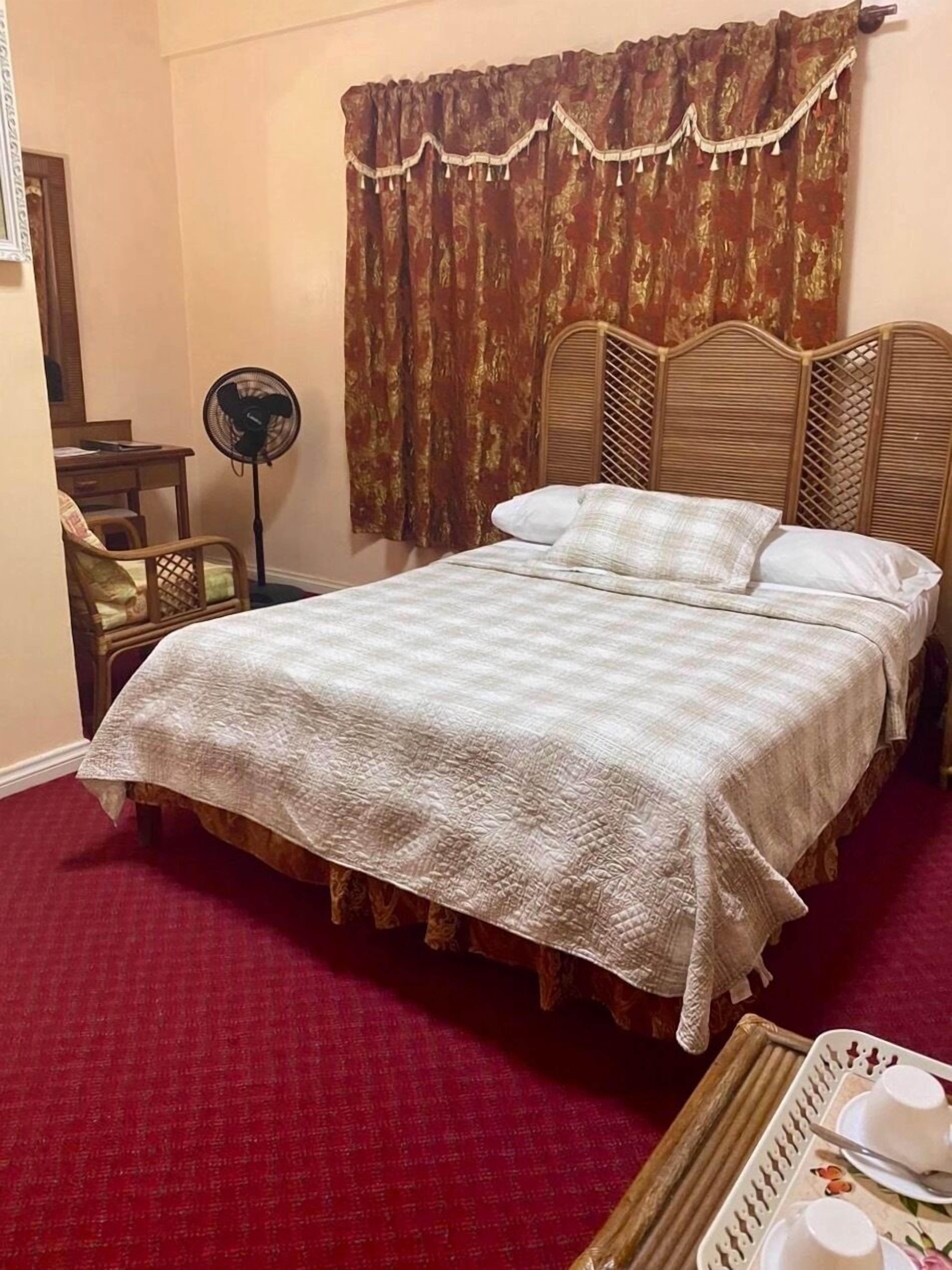 A hotel room with a bed, a wooden headboard, a red carpet, a fan, and a tray with cups and saucers.