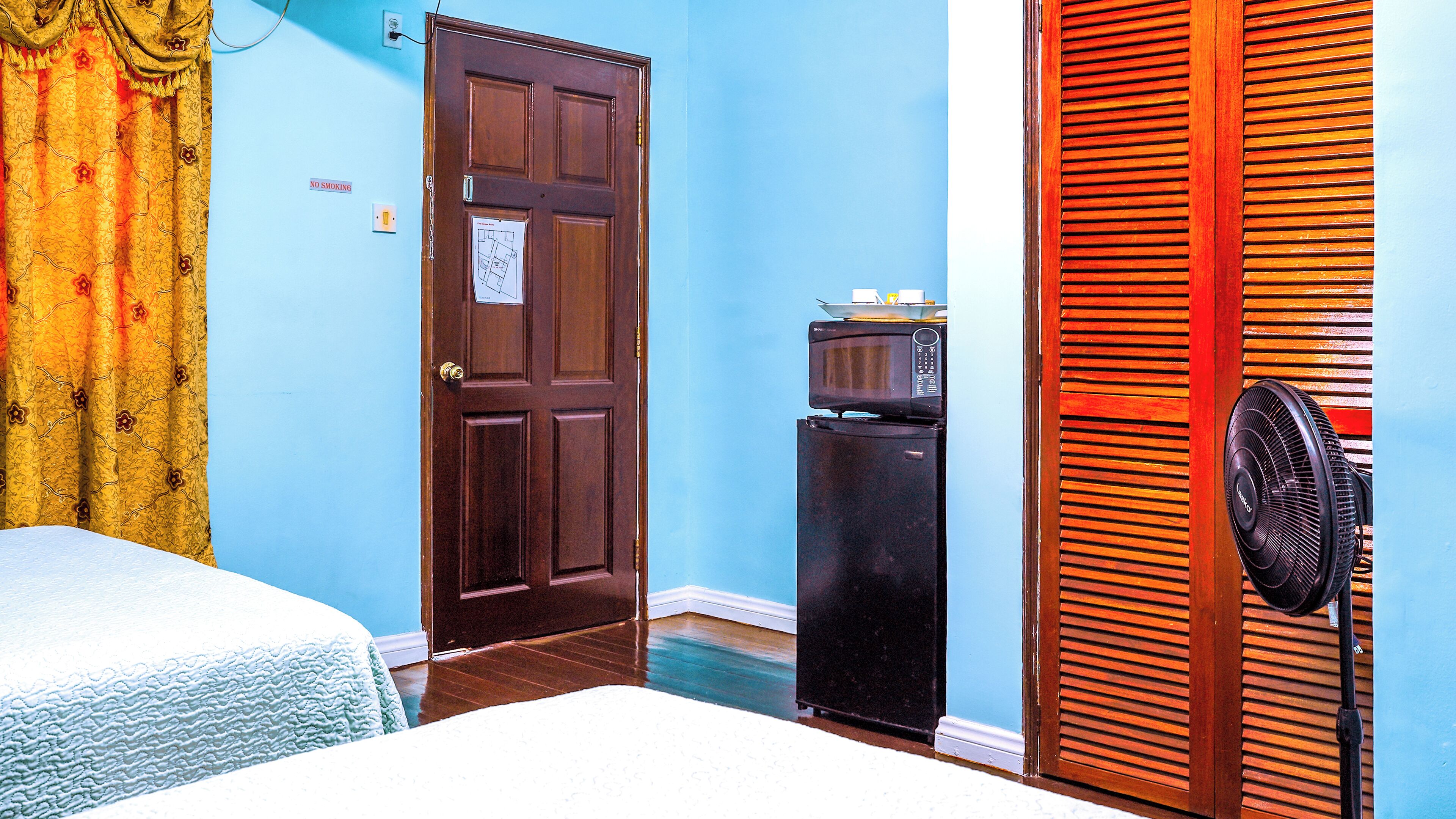 A room with a wooden door, a refrigerator, a fan, and a bed.