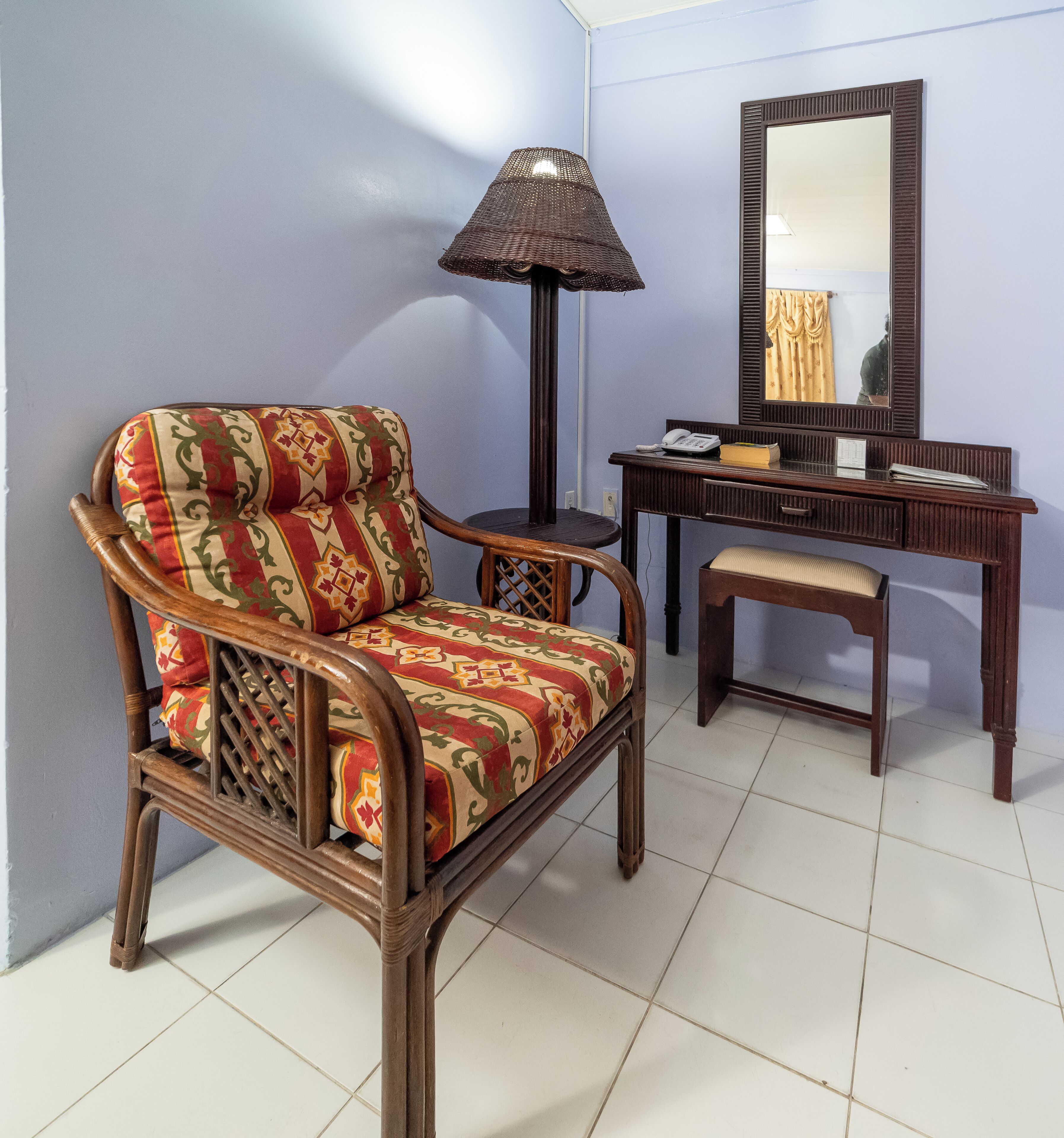 A room with a wooden chair with a patterned cushion, a small table, a lamp, a mirror, and a desk with a telephone and a book.