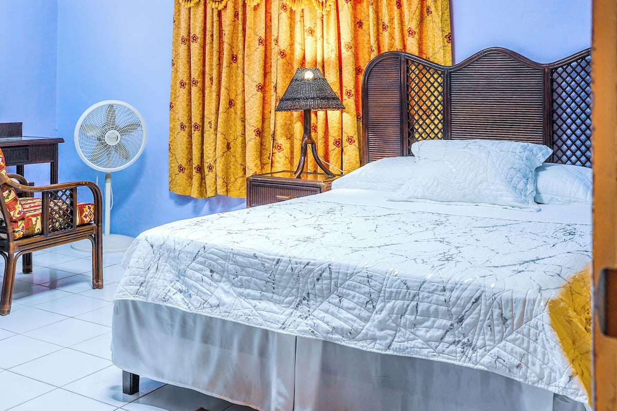 A bedroom with a bed, a chair, a fan, a lamp, and curtains.
