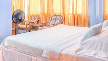 A hotel room with a bed, two chairs, a standing fan, and curtains.
