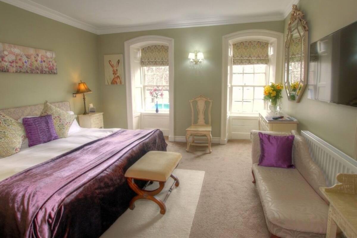 A bedroom with a bed, a chair, a bench, a sofa, a lamp, a mirror, and a TV.