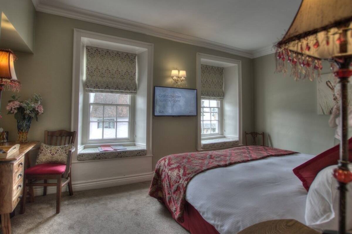 A bedroom with a bed, a desk, and two windows with decorative curtains.