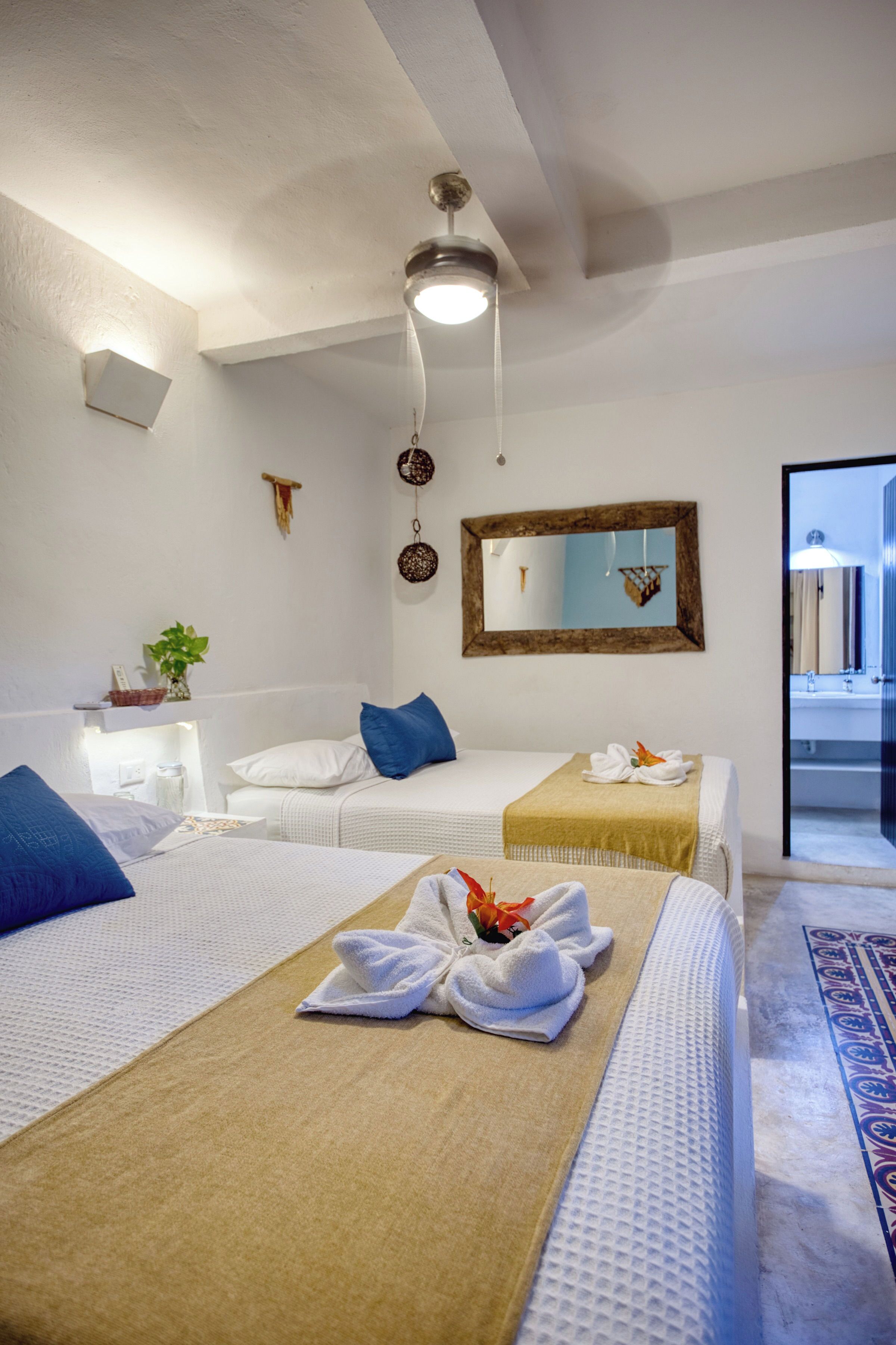 Habitació d'hotel amb dos llits, un bany visible per una porta oberta, un ventilador de sostre i un mirall fixat a la paret.