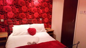 A hotel room with a bed, white bedding, a red heart-shaped pillow, and a wall covered in red roses. - The Fame (Blackpool)