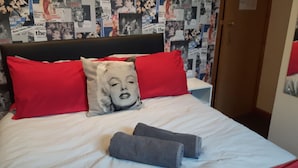 A bed with red and white pillows, a grey blanket, and a pillow with a pop culture print. - The Fame (Blackpool)