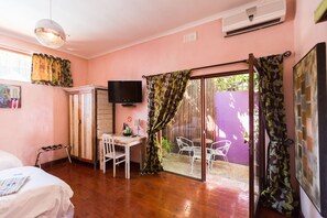 Une chambre comprenant un lit, un bureau, une chaise, un téléviseur et une porte-fenêtre coulissante donnant sur un espace extérieur aménagé avec des chaises.