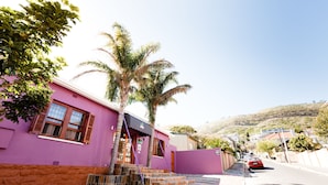 A pink building with a staircase leading to the entrance, palm trees, and a street with cars. - Sweet Olive Guesthouse (Cape Town)