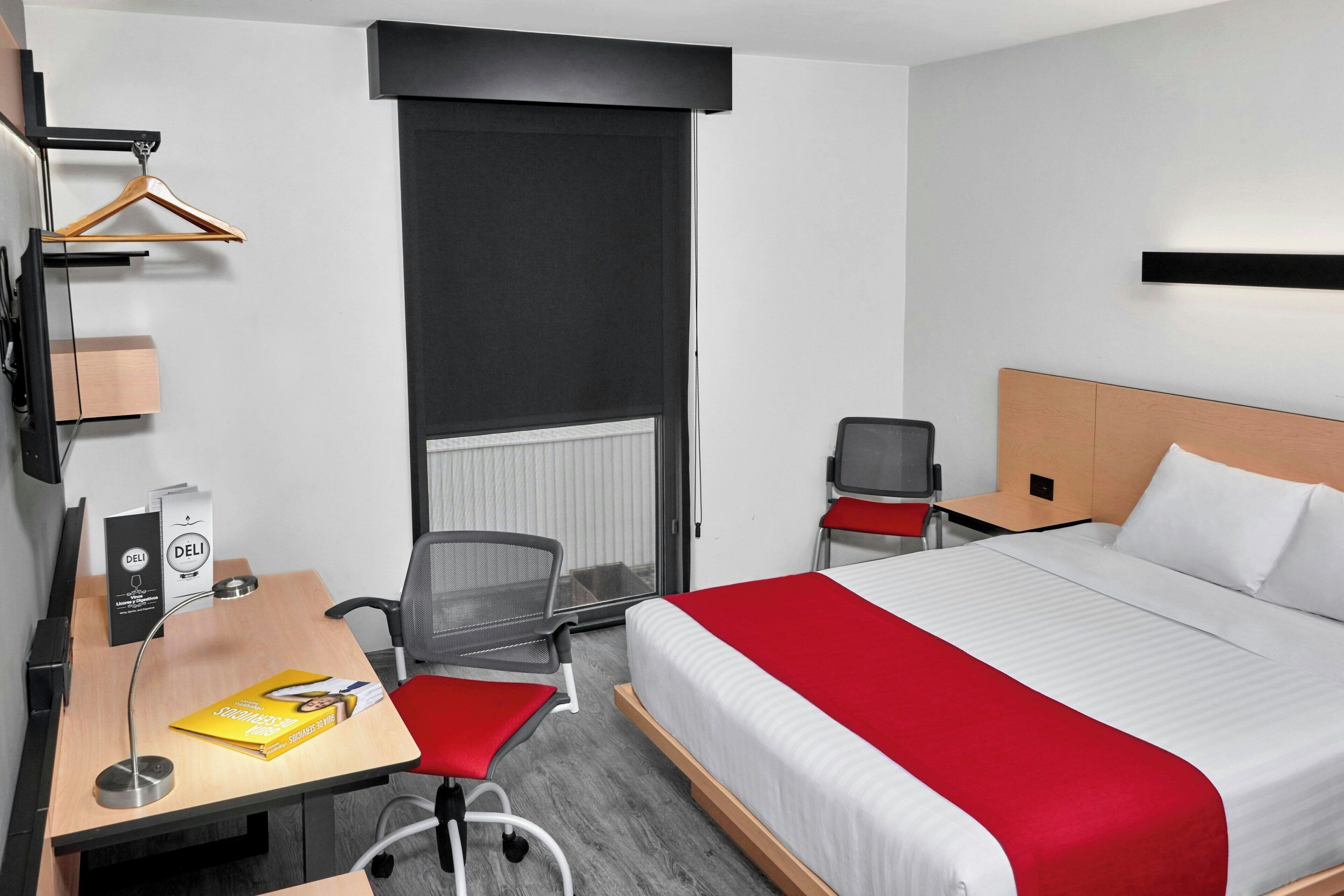 A hotel room with a bed, a desk with a chair, a hanging coat rack, and a radiator.