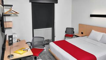 A hotel room with a bed, a desk with a chair, a hanging coat rack, and a radiator.