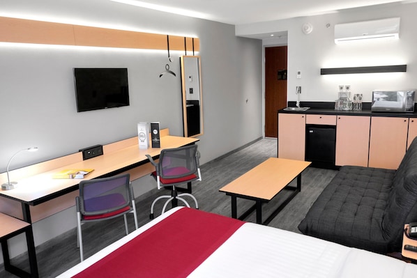 Una habitación de hotel moderna con cama, escritorio, silla, sofá, televisor, espejo, kitchenette y baño.