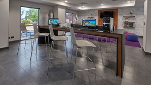 A modern office space with a long table, four white chairs, two desktop computers, a coffee machine, and a brick wall with shelves.