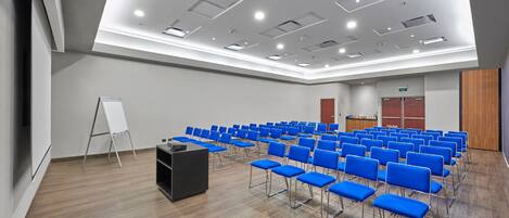 Un salón de conferencias con filas de sillas azules, un atril negro y una pizarra.