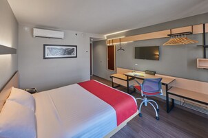 Habitación de hotel con cama, escritorio, silla y televisor en la pared.