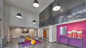 A spacious lobby with a modern design, featuring a central seating area with a sofa and chairs, a purple accent wall with artwork, and a glass railing overlooking the space.