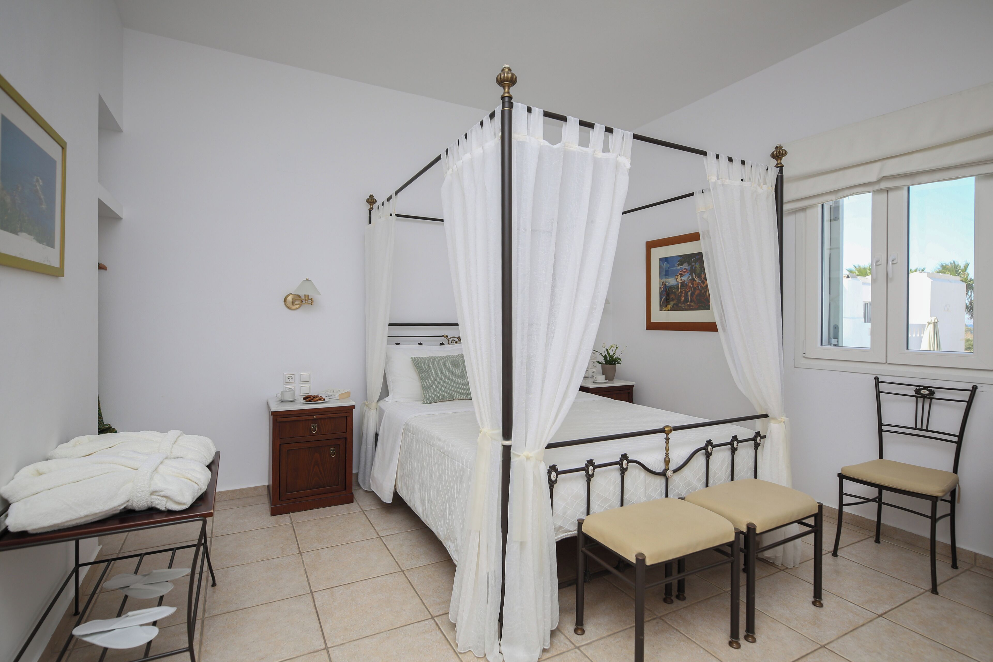 Un dormitorio con cama con dosel, una mesita de noche, una silla, un armario pequeño y una ventana con vistas a edificios.