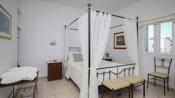 Un dormitorio con cama con dosel, una mesita de noche, una silla, un armario pequeño y una ventana con vistas a edificios.