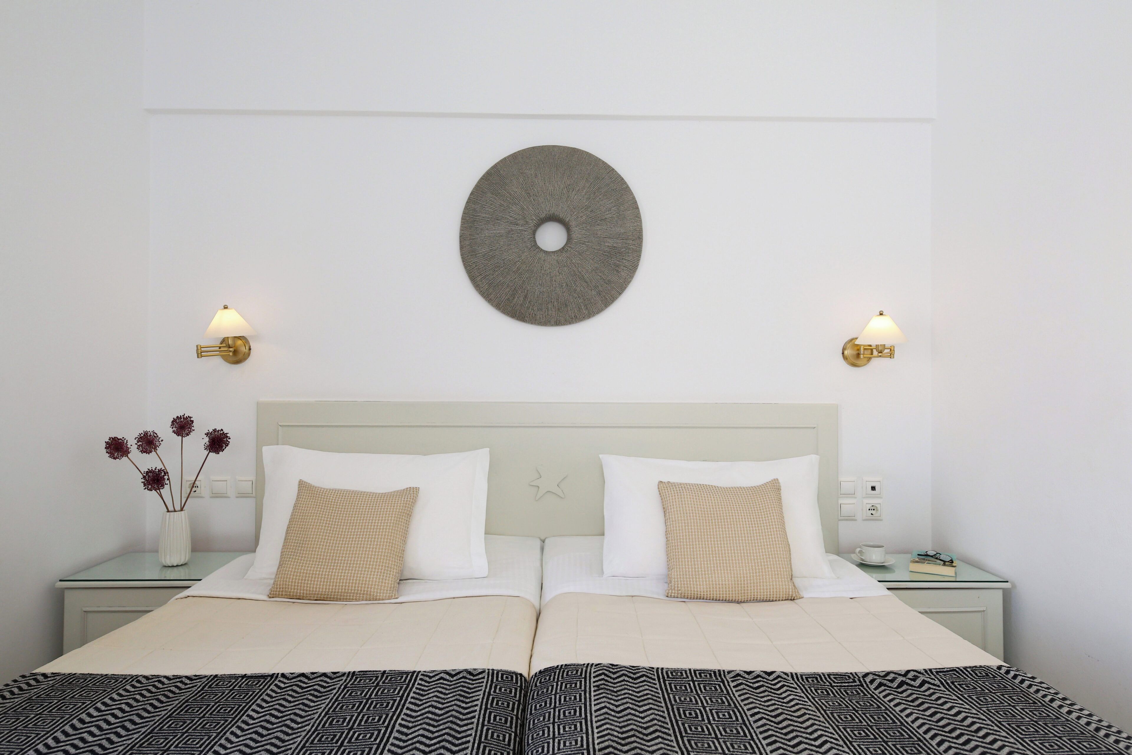 Una cama doble bien hecha con una colcha estampada, dos mesitas de noche con lámparas, un adorno circular en la pared y un jarrón con flores en la mesita de noche.