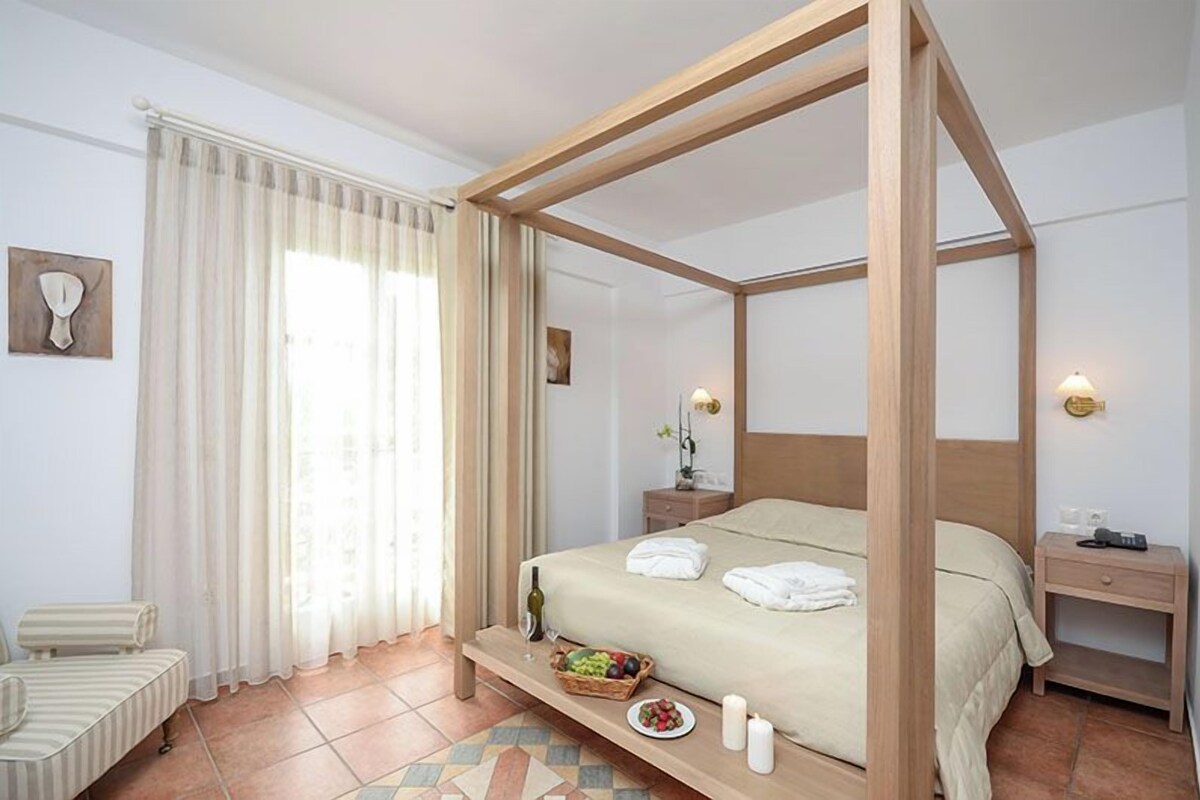 A four-poster bed with a wooden frame, a bedside table with a lamp, a small bedside table with a phone, a bedside table with a vase, and a small table with a fruit bowl and candles.