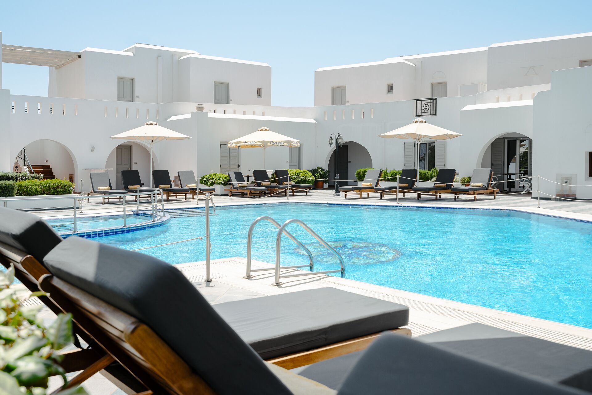 A pool area with lounge chairs and umbrellas, surrounded by white buildings with arched doorways.