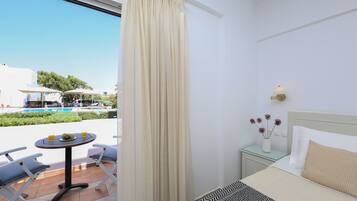A hotel room with a bed, a bedside table with a lamp, a small table with a fruit bowl and drinks, and a view of a pool and greenery outside.