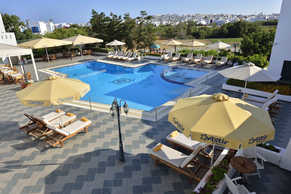 A pool area with sun loungers and umbrellas, surrounded by a tiled deck and overlooking a cityscape.