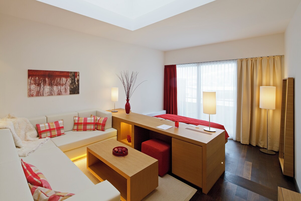 A modern hotel room with a sofa, a small desk, a red ottoman, a vase, and a painting on the wall.