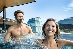 A couple in a hot tub with a modern glass building in the background. - TAUERN SPA Zell am See - Kaprun (Kaprun)