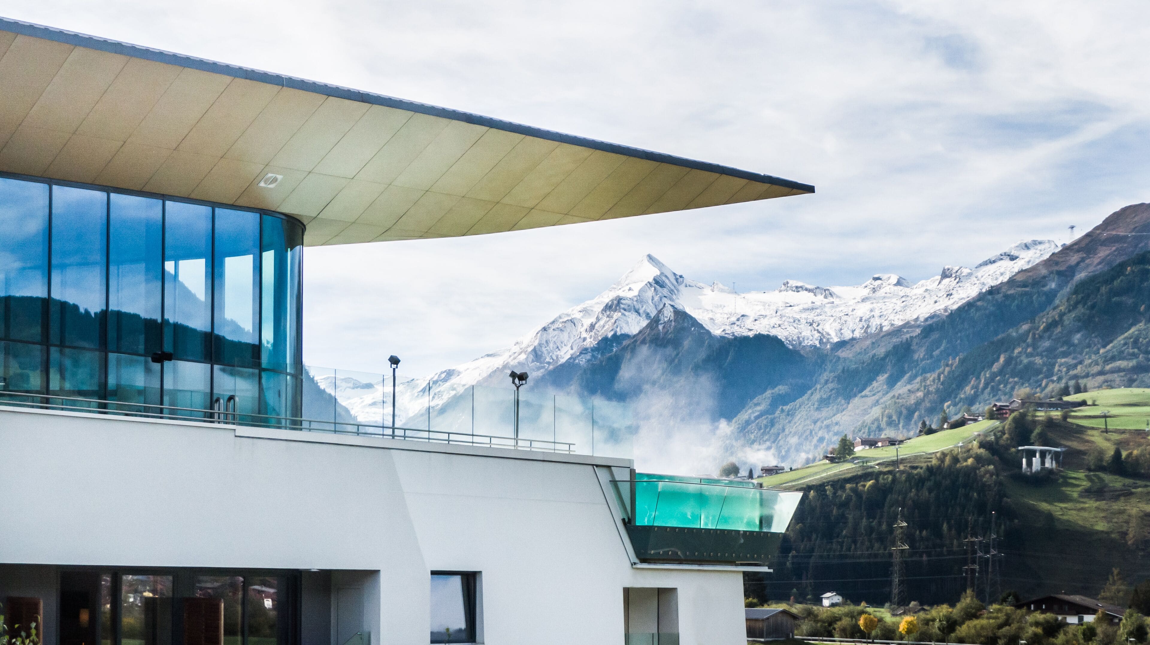 A modern building with large glass windows and a flat roof, overlooking a mountainous landscape with snow-capped peaks.