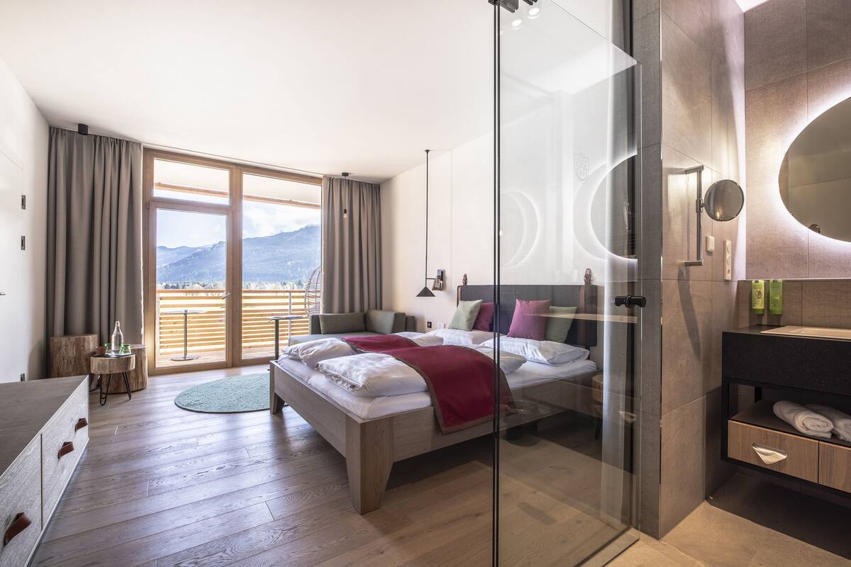 Ein modernes Schlafzimmer mit einem großen Bett, einer Dusche mit Glastrennwand, Holzboden und Bergblick durch das Fenster.