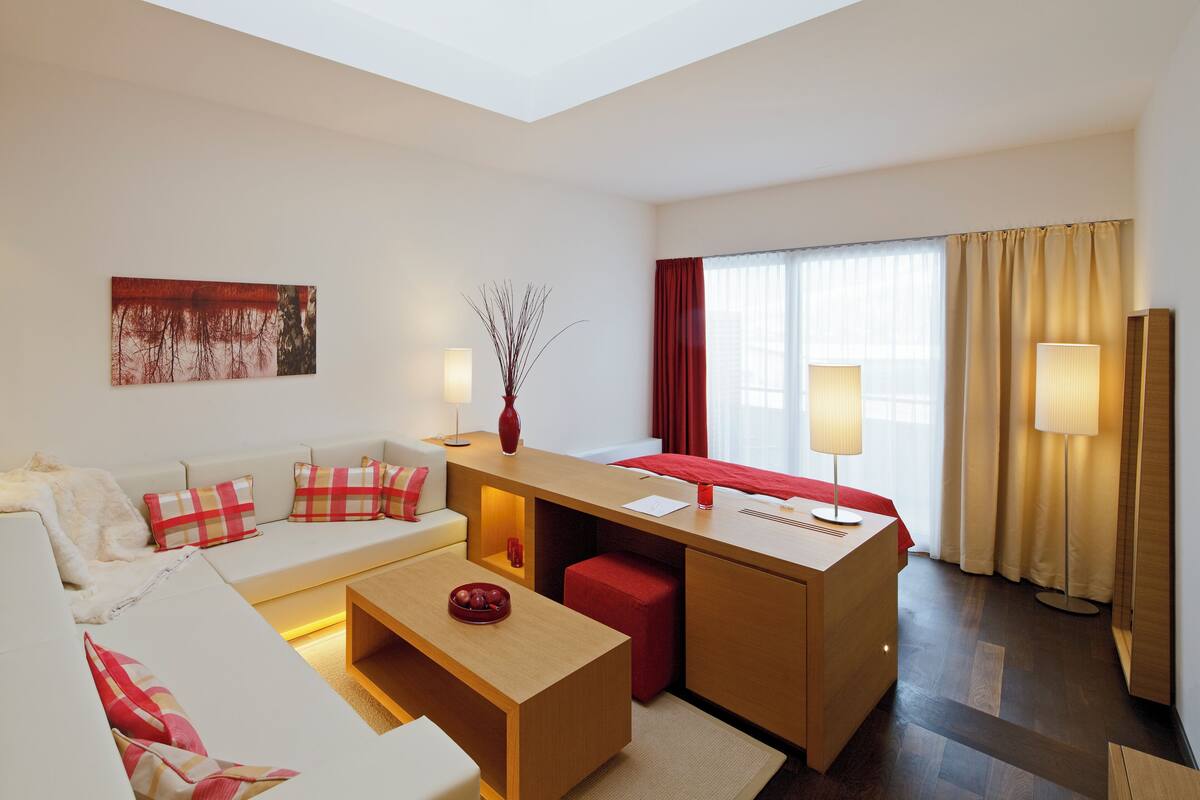 Ein modernes Hotelzimmer mit einer Couch, einem kleinen Schreibtisch, einem roten Hocker, einer Vase und einem Bild an der Wand.