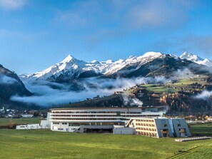 A modern building with a flat roof and multiple windows, surrounded by green fields and mountains in the background. - TAUERN SPA Zell am See - Kaprun (Kaprun)
