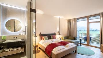 A modern hotel room with a large bed, a sitting area, a balcony with a view, and a bathroom with a mirror and sink.