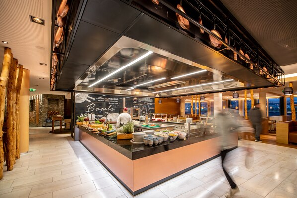 Un restaurante moderno con una isla central que exhibe comida y bebidas, un menú en pizarra y vistas a la ciudad.