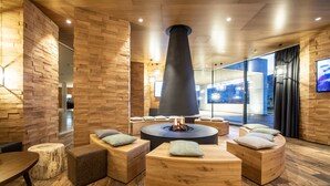 A modern lounge area with a central fire feature, wooden seating, and a large window offering a view. - TAUERN SPA Zell am See - Kaprun (Kaprun)