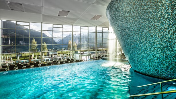 An indoor swimming pool with clear blue water, large windows offering mountain views, and a unique textured wall. - TAUERN SPA Zell am See - Kaprun (Kaprun)