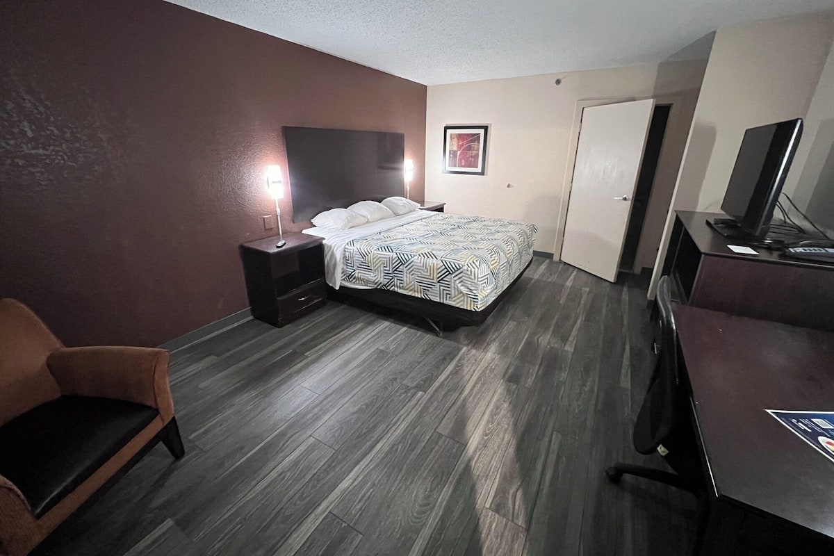 Une chambre d’hôtel comprenant un lit, un bureau avec un ordinateur, une chaise et une table de chevet avec une lampe.