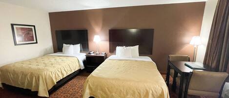 A hotel room with two beds, a desk, a chair, and a nightstand with a lamp.