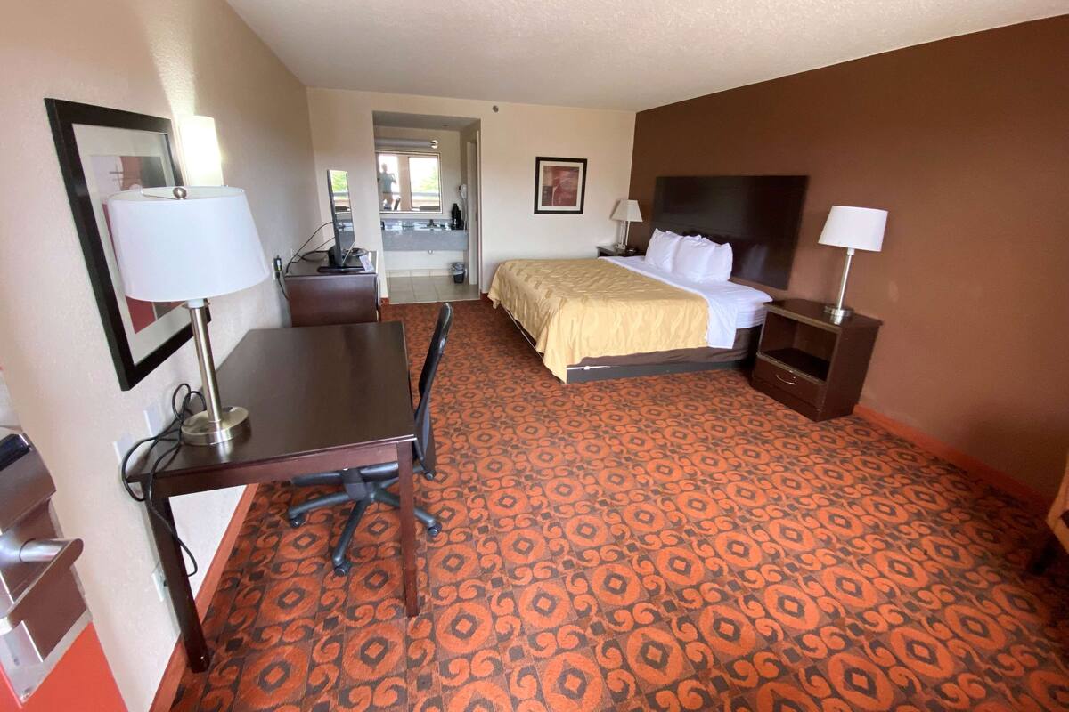 Une chambre d’hôtel avec un lit, un bureau, une chaise et une salle de bain visible à travers une porte ouverte.
