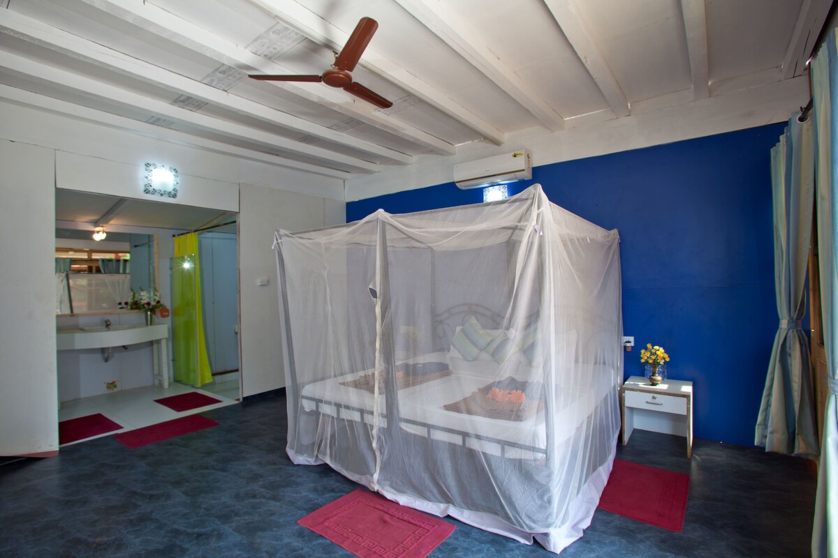 Ein Zimmer mit einem Bett unter einem Moskitonetz, einem Nachttisch mit Blumenvase und einer blauen Wand.