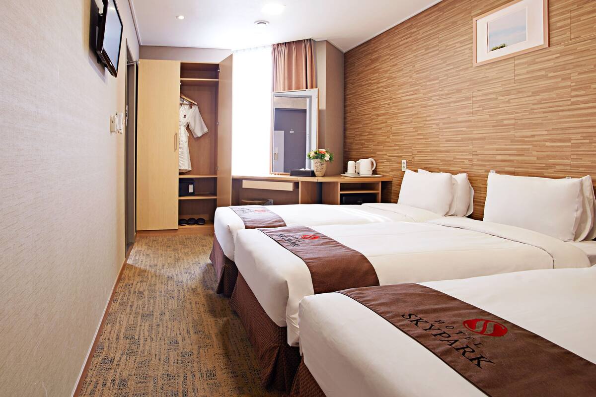 A hotel room with two beds, a wooden headboard, a desk with a chair, a wardrobe, and a window with curtains.