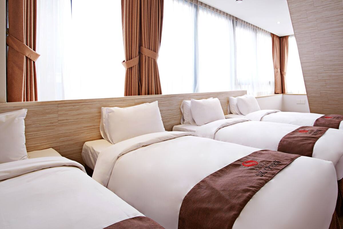 A hotel room with four beds arranged in a row, a wooden headboard, and large windows with curtains.