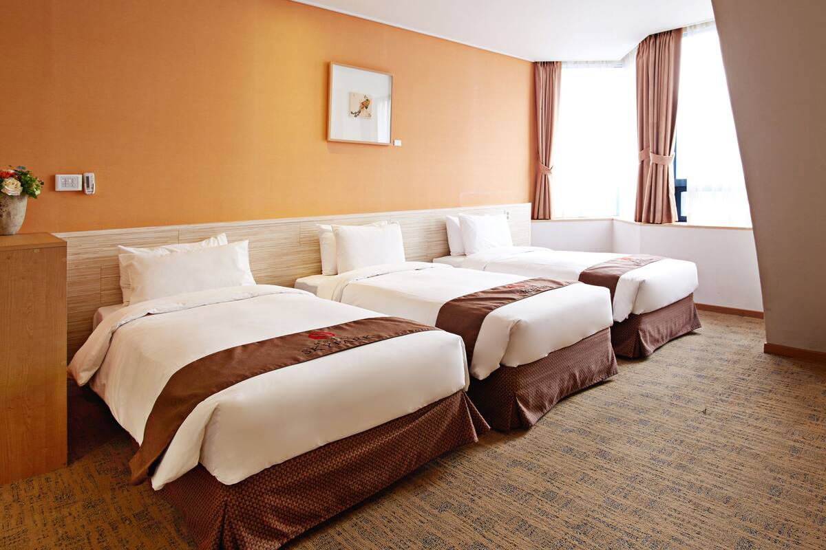 A hotel room with three beds, a wooden headboard, a framed picture on the wall, and a window with curtains.